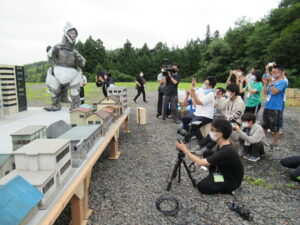 屋外の砂利敷きの会場で、精巧なミニチュアの街並みの奥に立つ怪獣の着ぐるみを、手前のカメラマンやスマートフォンを構えた大勢の塾生たちが一斉に撮影している写真
