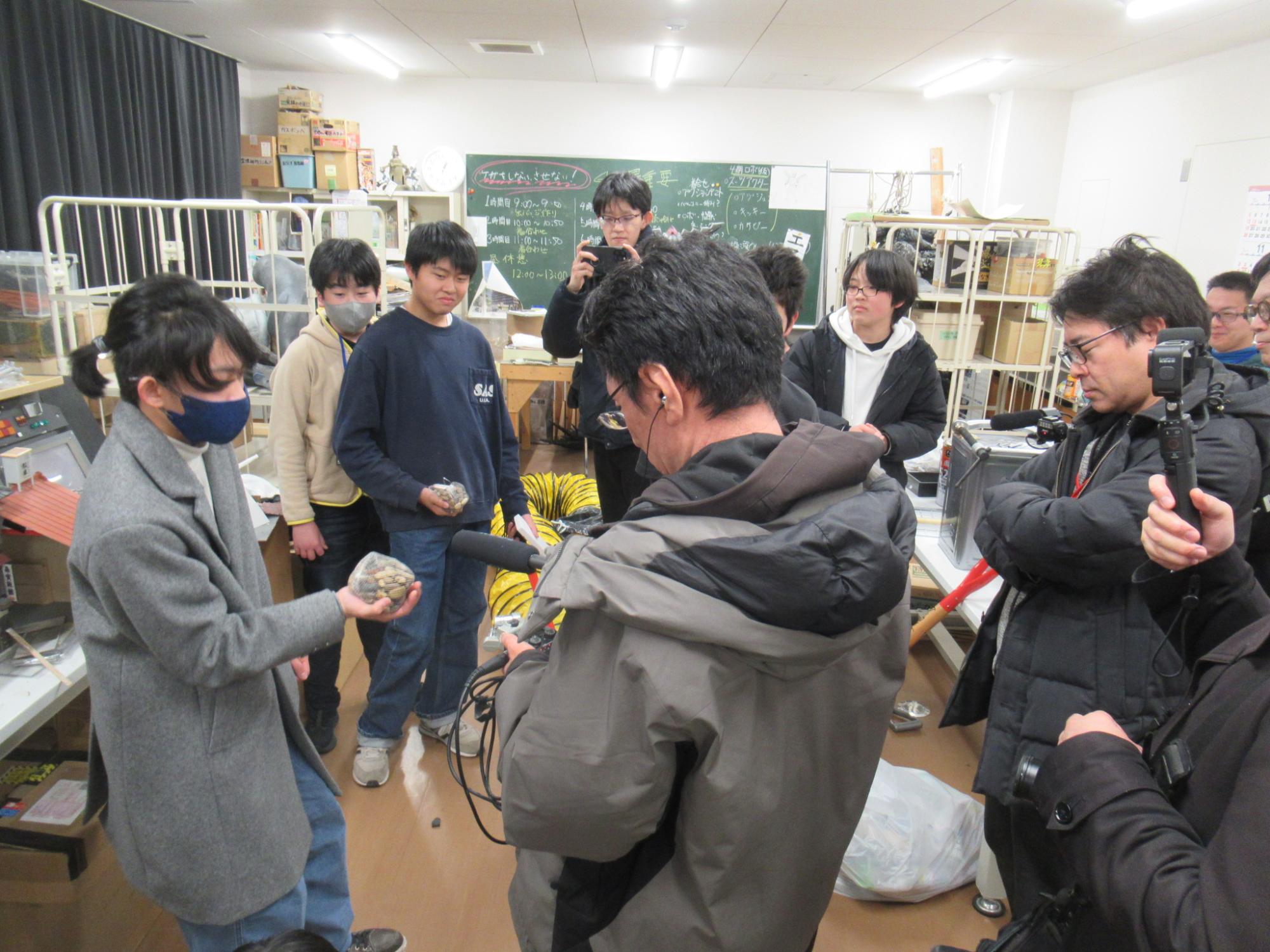 多様なものが置いてある作業室で効果音の元の音になるデータを収録している様子、小石が複数入ったネットをゆすって音を出す塾生とマイクで録音している講師がいる
