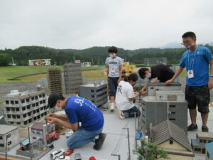 屋外の砂利敷きの広場で、塾生が精巧に作られたビルや民家のミニチュア模型が並ぶ撮影用セットが置かれ、電柱や車両などの細かなパーツを慎重に配置して街並みを調整している写真
