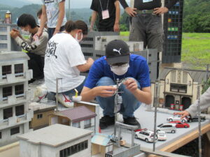 屋外の砂利敷きの広場で、ビルや民家のミニチュア模型が並ぶ撮影用セットの上に乗り、電柱や車両などの細かなパーツを慎重に配置して街並みを調整している、数名の塾生たちの写真