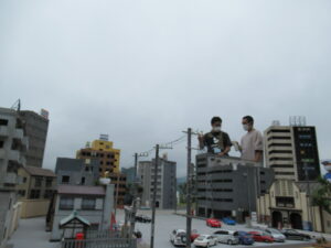 ビルや民家のミニチュア模型が並ぶ撮影用セットの後ろで2人の男性が並んで話をいる様子の写真