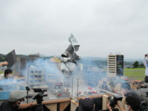 ビルや民家のミニチュア模型が並ぶ撮影用セットの間に煙が立ち込め、中央に立つ怪獣の着ぐるみを撮影している写真