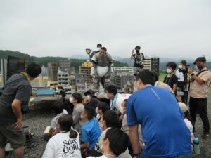 屋外の砂利敷きの広場で、ミニチュアの街並みが広がる撮影用セットの中央に、上半身を脱いだ状態の怪獣の着ぐるみを着た男性が立ち、その様子を大勢の塾生や講師が取り囲んで見つめている写真