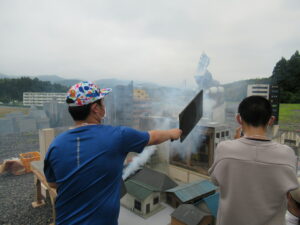 ミニチュアの街並みが広がる撮影用セットに白煙を漂わせ、スタッフがうちわで扇いで煙の動きを調節しているのを塾生が隣で見ている写真