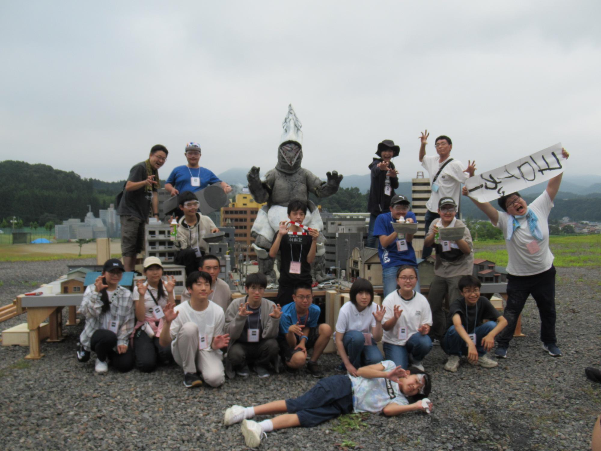 屋外の砂利の上で、ミニチュアの街並みを背景に怪獣の着ぐるみを中心に囲み、手で爪を立てるポーズをしたり「ヨロイガー」と書かれた手書きの旗を掲げたりした大勢の塾生たちと講師の集合写真