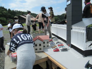 晴天の屋外で、並べられた多くのミニカーや民家、そして巨大な黒い塔のような模型を囲み、塾生が配置の微調整を行ったりしている写真