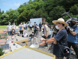 晴天の屋外で、麦わら帽子を被った講師がスプレーを使って特殊効果を施し、その様子を大勢の塾生が興味深そうに見つめている写真