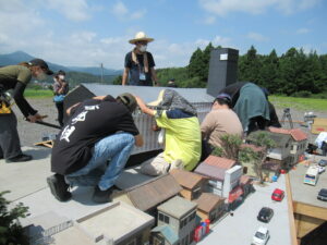 青空の下でタオルや帽子を被った複数の塾生が、民家や車が並ぶ精密なミニチュアの街並みの中に身をかがめ、大きな建物の模型を慎重に設置している様子の写真