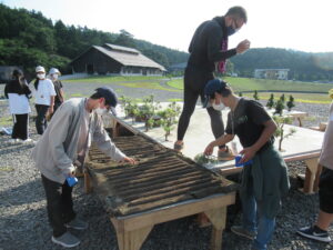 屋外で、数名の塾生が木製の台座の上に並べられた樹木のミニチュア模型を丁寧に整えている様子の写真