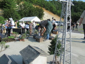 屋外にて、鉄塔や民家の模型が並ぶミニチュアの街並みの中で、塾生が腰をかがめて建物や樹木の配置を細かく調整している写真