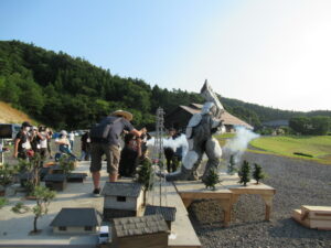 屋外にて、怪獣の着ぐるみが白い煙を上げながらミニチュアの街並みに立ちはだかるシーンを、カメラマたちが低いアングルから撮影をしているのを塾生が後ろから見つめている写真