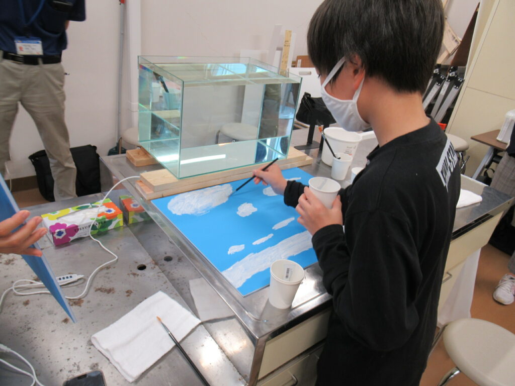 明るい室内で、白いマスクをつけた少年が机に向かい、青い画用紙に白い絵の具を使って雲のような模様を丁寧に描き込んでいる様子を作業中の少年の背後から捉えた、絵画教室での熱心な制作風景の写真