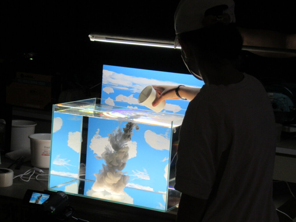 暗い室内で、青空と白い雲が描かれたパネルを背景にした透明な水槽の中に、白い液体がゆっくりと注がれ、まるで本物の雲が湧き上がるような幻想的な視覚効果を生み出している瞬間を捉えた、特殊な美術制作の風景の写真