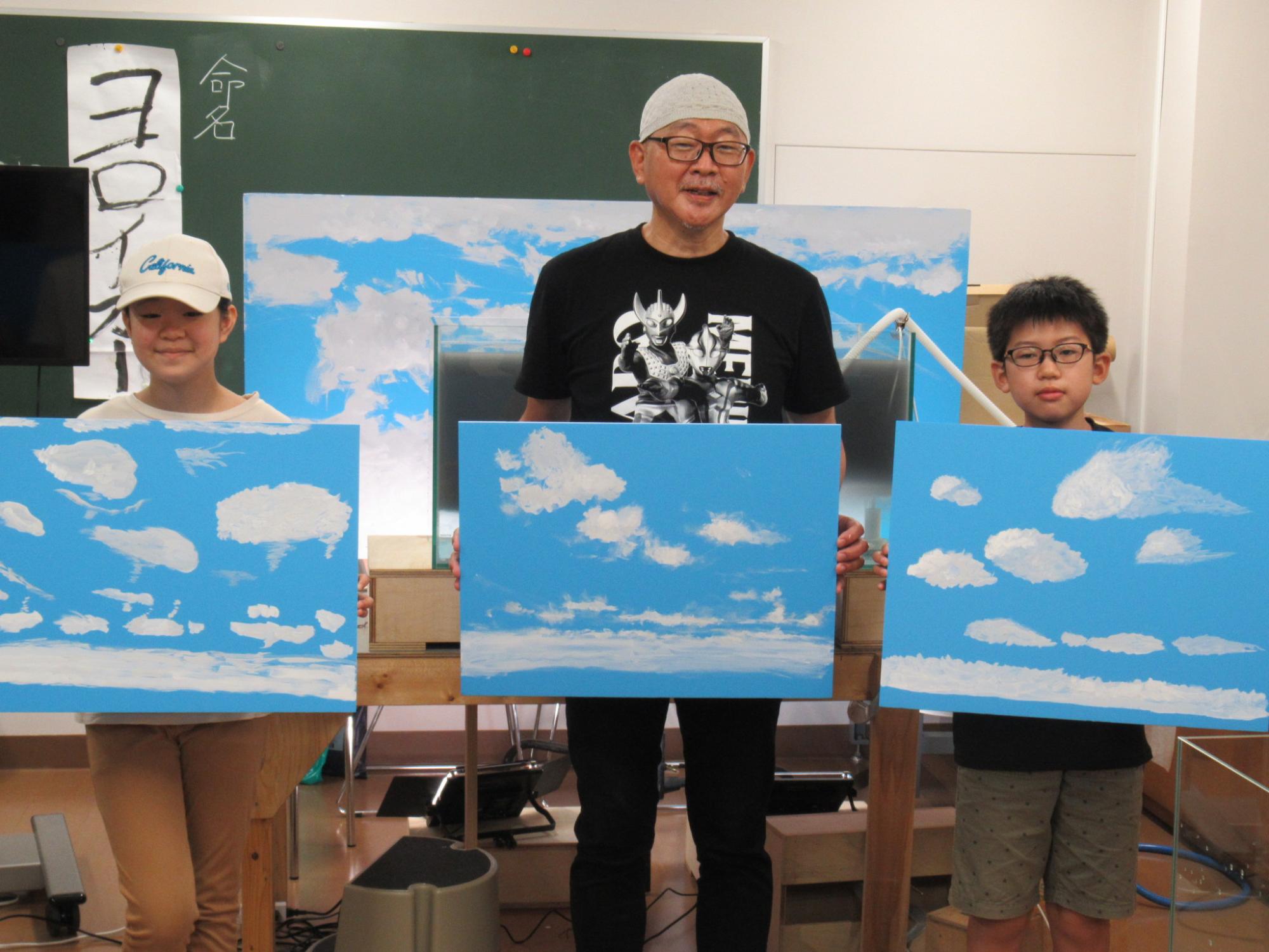 明るい室内で、黒いTシャツを着て眼鏡をかけた年配の男性と、帽子を被った少女、そして眼鏡をかけた少年が、青空と白い雲を描いた三枚の鮮やかな絵画をそれぞれ手に持って並んでいる、創作活動の成果を披露する記念写真