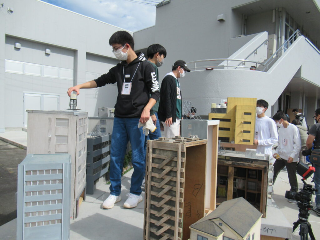 屋外で塾生たちがミニチュアのビルや家屋が並ぶジオラマセットの設営を行っており、手前の塾生がビルの屋上にパーツを慎重に配置している写真