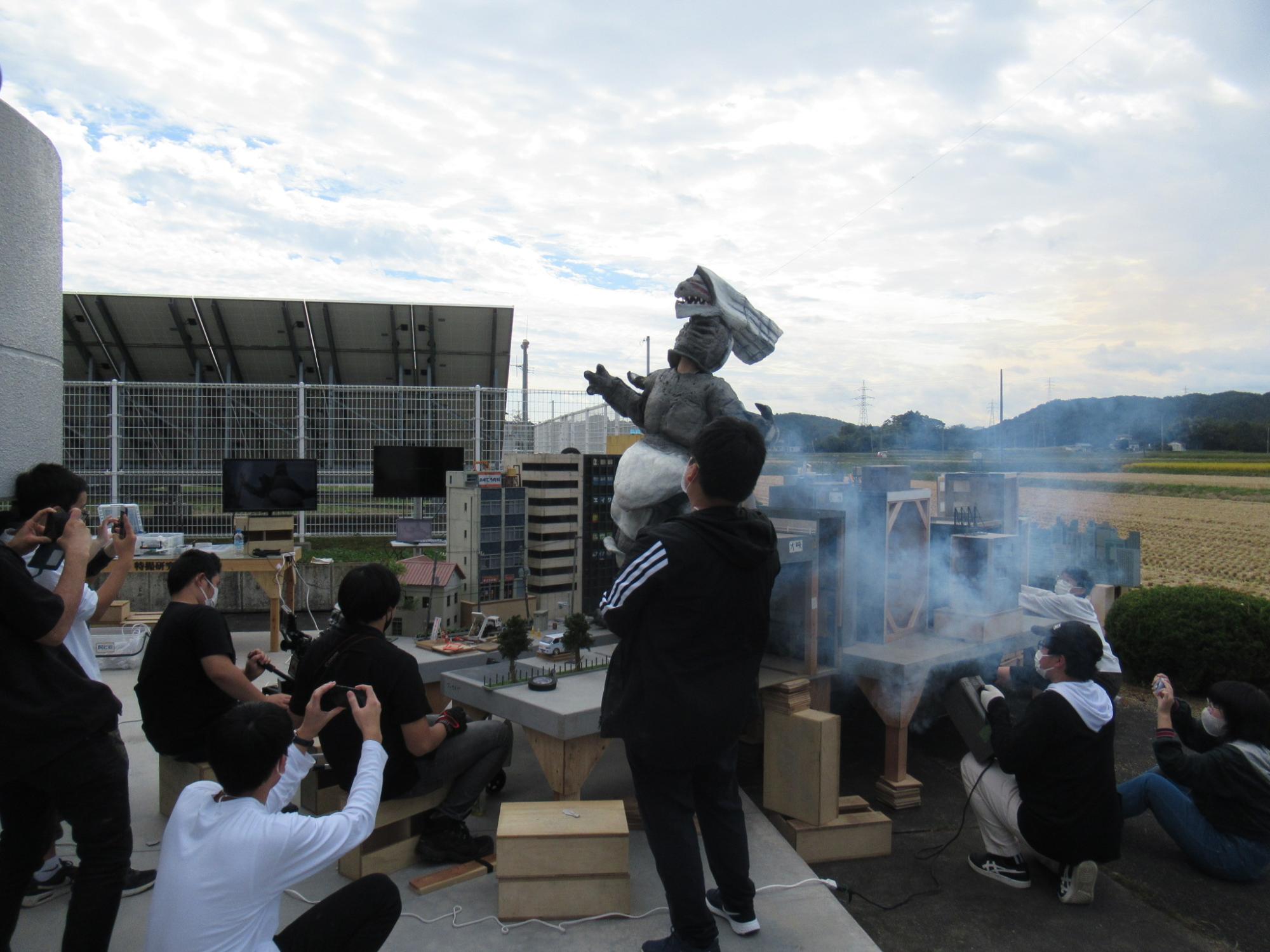 屋外でミニチュアのビル群で煙が立ち込めるジオラマのセットを囲み、怪獣の着ぐるみを操る演者や、その様子をスマートフォンやカメラで撮影する塾生たちの写真