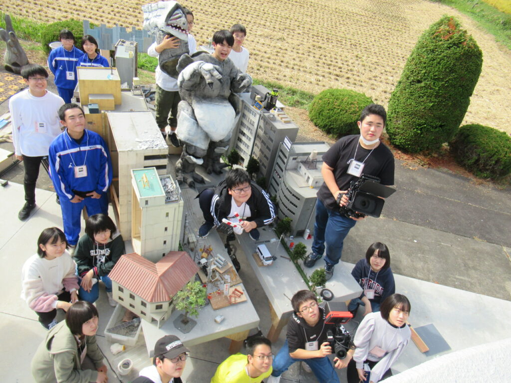 屋外で怪獣の着ぐるみやビル群の精巧なミニチュアセット上で、カメラやカチンコを手にした多くの塾生たちが集まって記念撮影を行っている写真