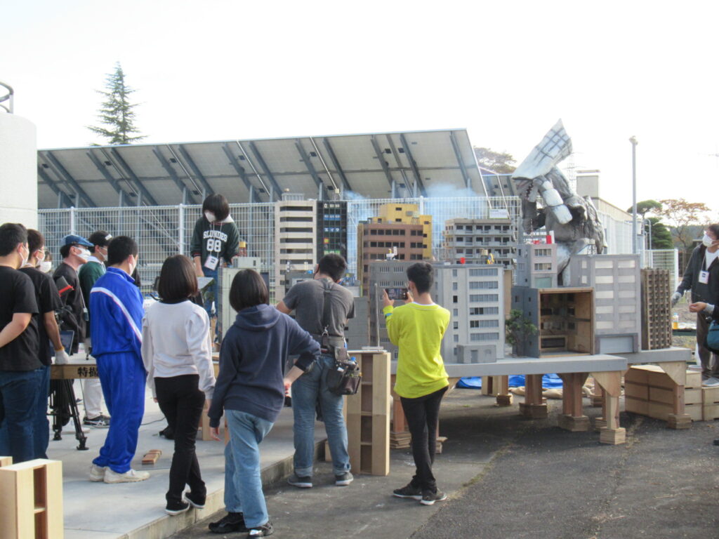 屋外でミニチュアのビル群に怪獣の着ぐるみが立ち、その様子を多くの塾生がスマートフォンやカメラで撮影している写真