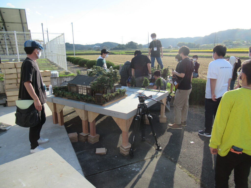 のどかな田園風景が広がる屋外で、多くの塾生が日本家屋のミニチュアセットを木製の台座に設置し、三脚に据えられたカメラを前にして撮影の準備や打ち合わせを行っている写真