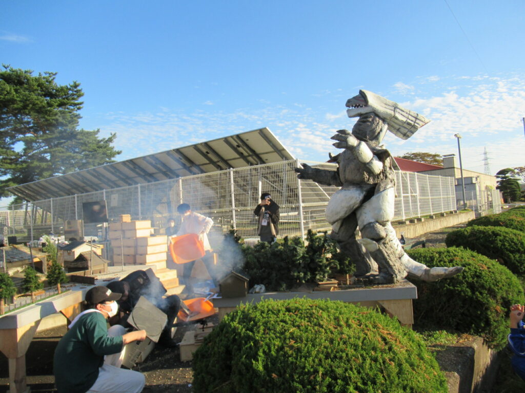 野外で大きな怪獣の着ぐるみを台座に立たせ、スタッフが周囲でスモークを焚き角度を調整したりして、特撮シーンの作り込みを行っている写真