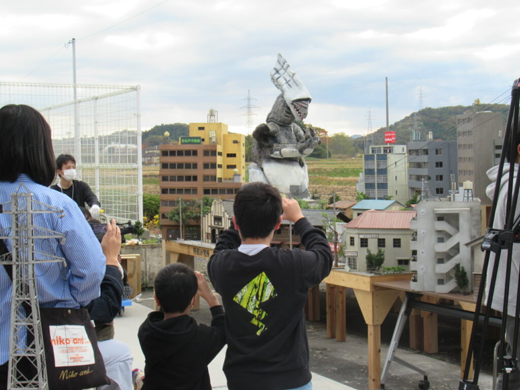屋外に組まれた精巧な都市模型の中央に巨大な怪獣模型が立ち上がり、それを多くの来場者が熱心に撮影している様子を捉えた写真