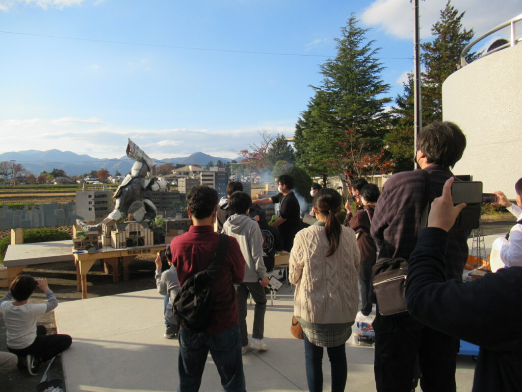 屋外で巨大な怪獣模型を囲む人々がミニチュア都市の精巧なセットを眺め撮影している様子の写真