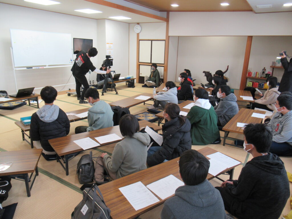 畳敷きの和室で、ホワイトボードやカメラ機材が設置された前方の講師を、塾生たちが資料を手にしながら向かい合わせになり、座卓に並んで座っている写真
