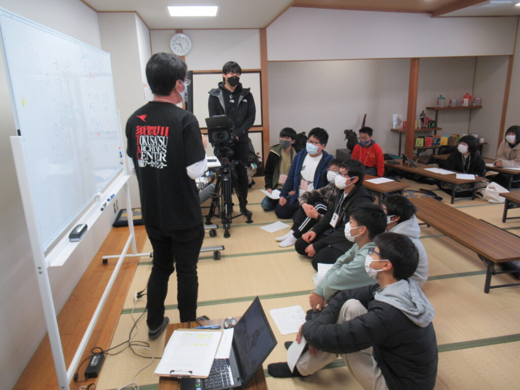 畳敷きの和室で、ホワイトボードの前に立つ講師が説明を行い、その様子をカメラで撮影するスタッフと、畳に座って話を聞き入る塾生たちの写真