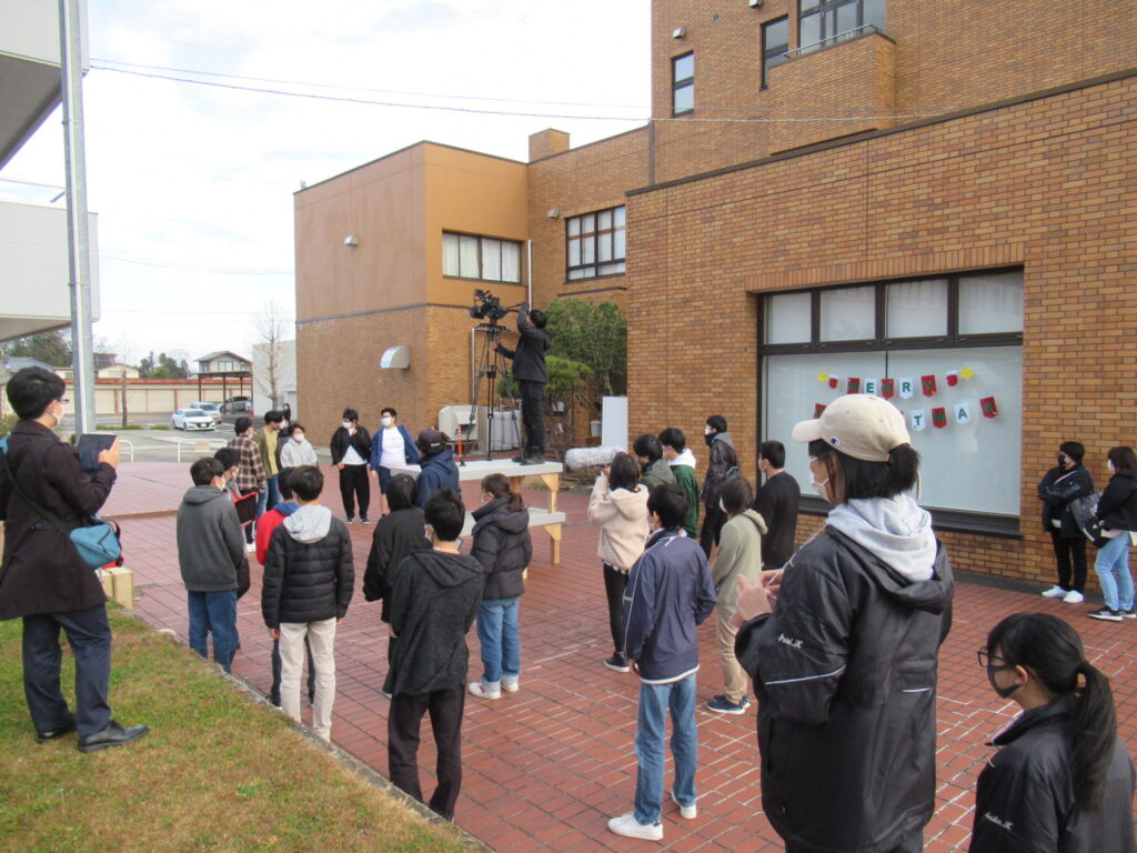 茶色のレンガ造りの建物の前の広場で、多くの塾生たちが集まり、台の上で三脚を立てて大きなカメラを構える撮影スタッフの写真