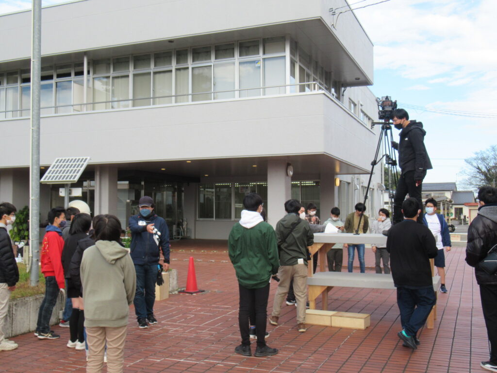 白い建物の前の広場で、台の上に三脚を立てて大きなカメラを構える撮影スタッフの様子を、多くの塾生たちが囲むように集まって見守っている写真