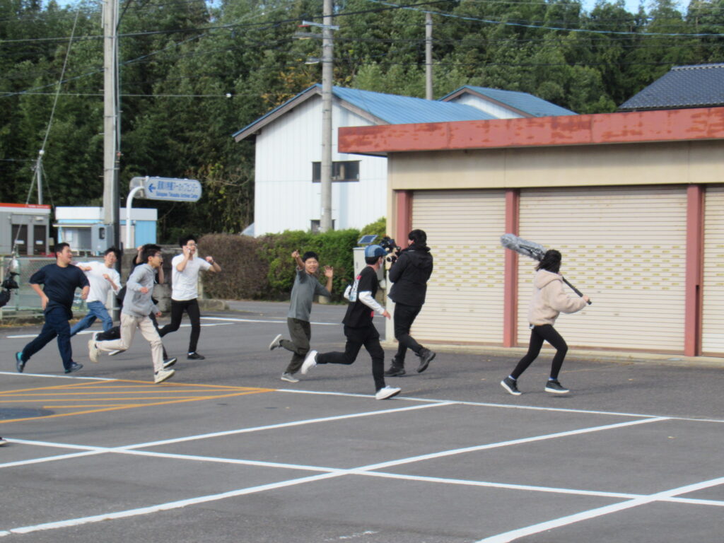 アスファルトが敷かれた広場で、機材を抱えて走る撮影スタッフを追いかけるように、多くの若者たちが全力で疾走している、映画のワンシーンを撮影している写真