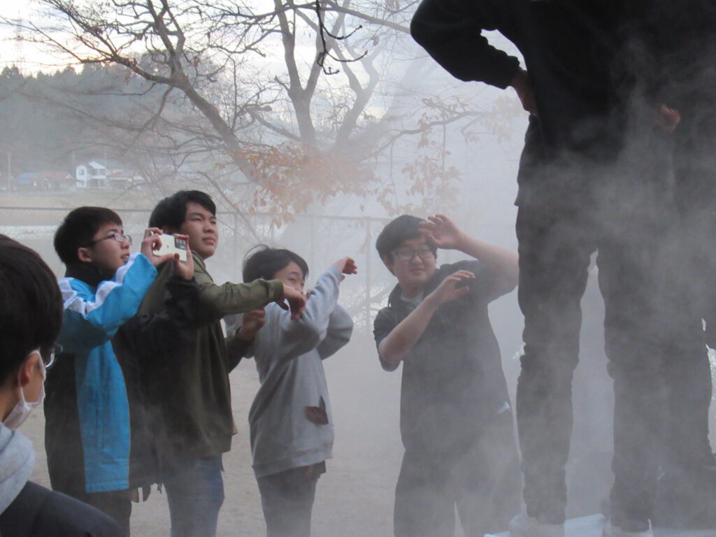 煙が立ち込める屋外で、前方に立つ数人の若者たちがスマートフォンのカメラを向けたり、驚いたような仕草で見つめたりしている写真