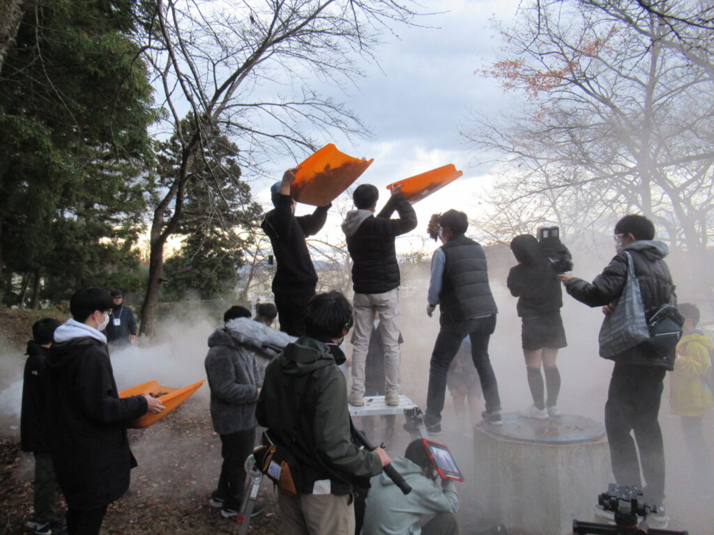 白煙が立ち込める森の中で、落ち葉が入ったオレンジ色の入れ物を高く掲げる塾生やカメラを構える撮影者、それを見守る塾生たちが集まり、特撮映画の特殊効果を用いた撮影が行われている写真