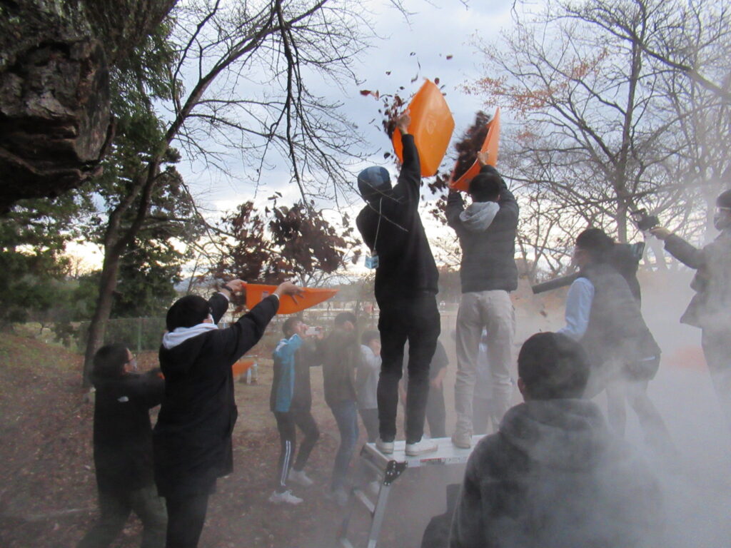 森の中で、脚立に登った塾生がオレンジ色の入れ物から枯れ葉を高く撒き散らし、周囲に白煙が漂う中で塾生たちがその光景を撮影したり見守ったり、演技をしている、特撮映画の演出シーンの写真