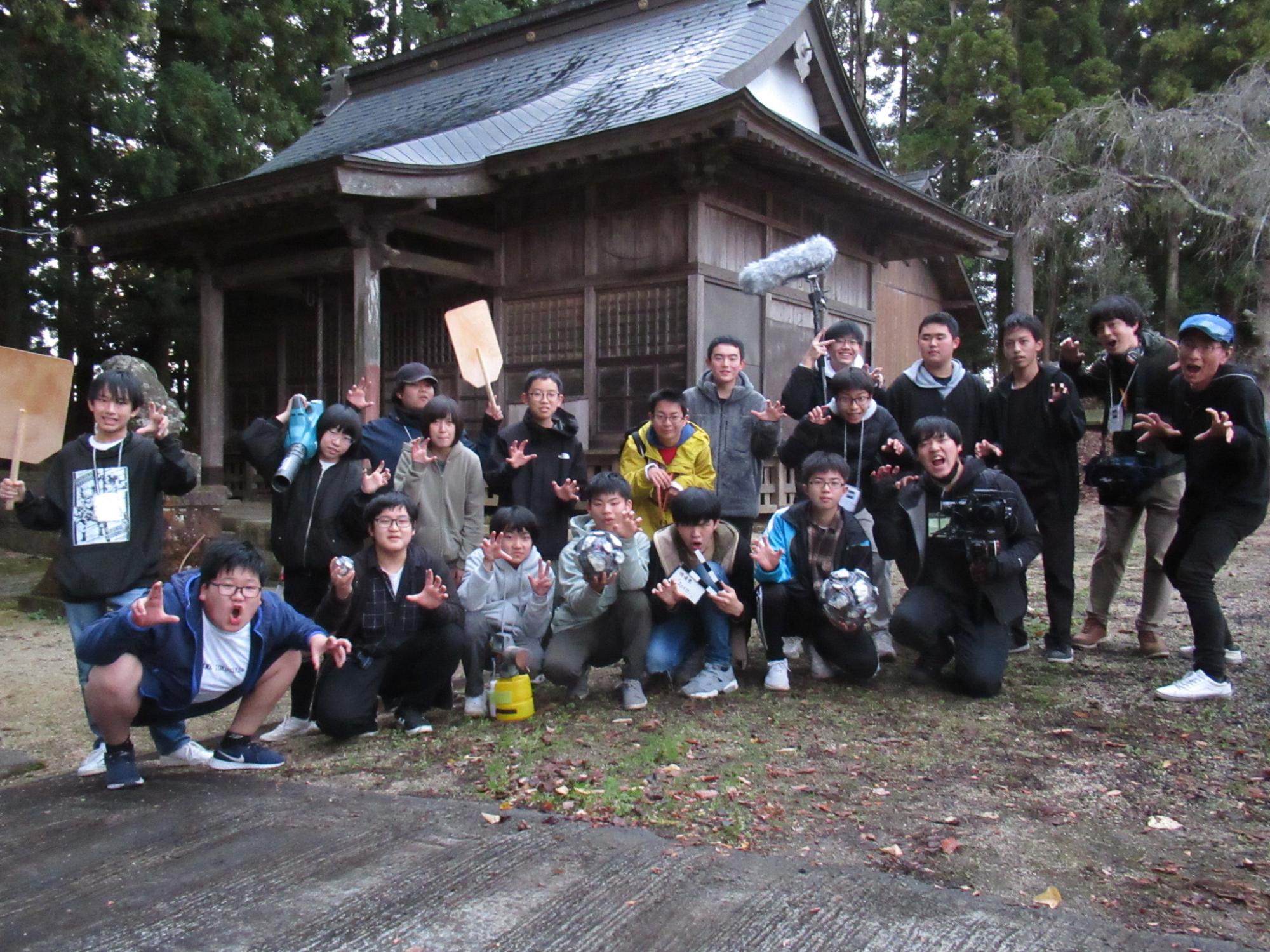 古い木造の神社の前で、多くの塾生が様々な撮影機材や小道具を手に持ち、屈んだりポーズをして写した集合写真