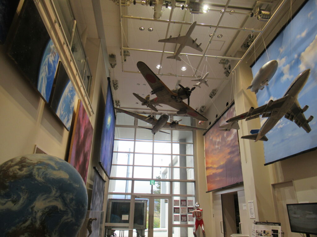 高い天井から零戦や旅客機の模型が吊り下げられ、壁面には地球や空の絵画が飾られた、特撮映画の資料や模型を展示する施設の内部を、手前に地球儀のような造形物を配置して奥行き感たっぷりに見渡した写真