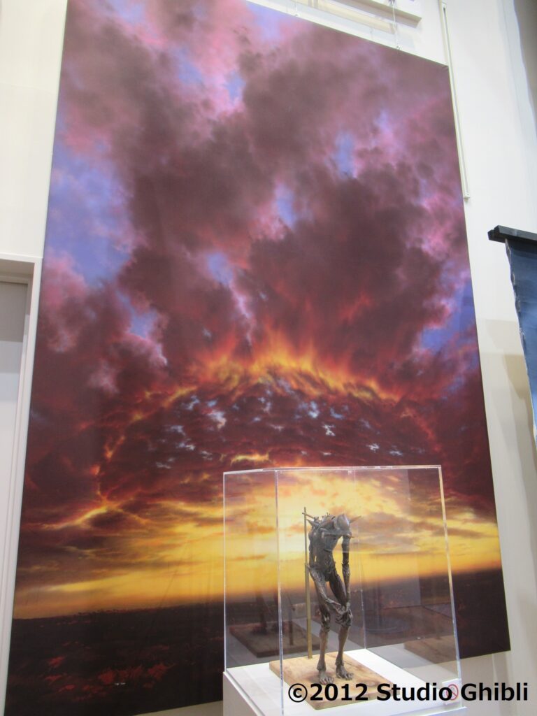 燃えるようなオレンジ色の空と、紫や黒の重厚な雲が劇的に広がる大空を描いた巨大な背景画を背に、透明なケースに入った痩身の巨神兵と思われるブロンズ色の彫像が展示されている写真