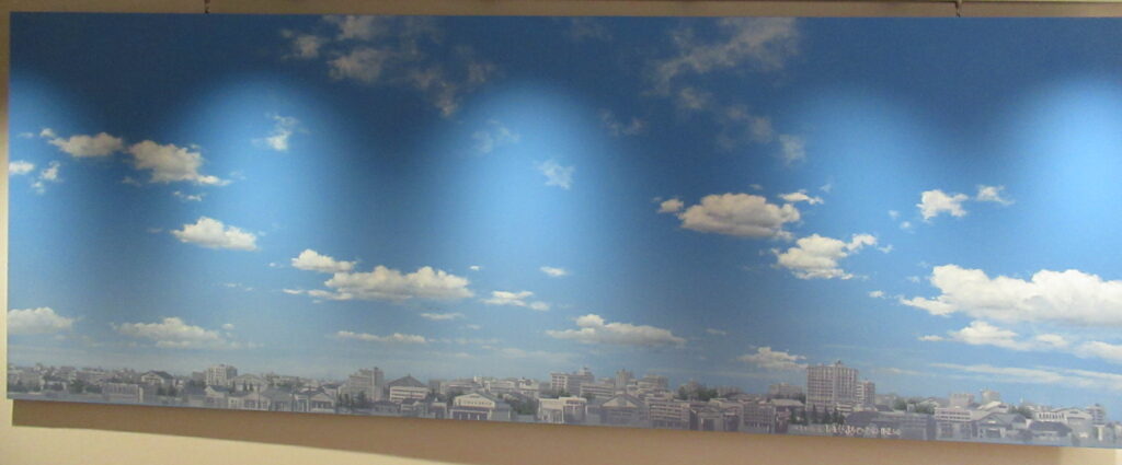 青空に白い雲が浮かび、画面下部にはどこまでも続く街並みが精緻に描かれた背景画