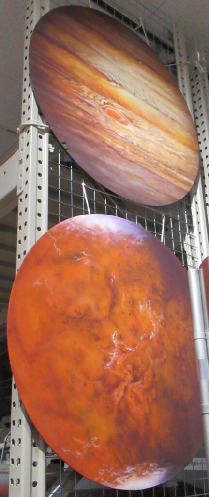 メタルラックに吊り下げられた、オレンジ色の斑点模様が広がる火星のような惑星と、大気の層や巨大な赤斑が緻密に描き込まれた木星のような惑星を描いた円形の背景画