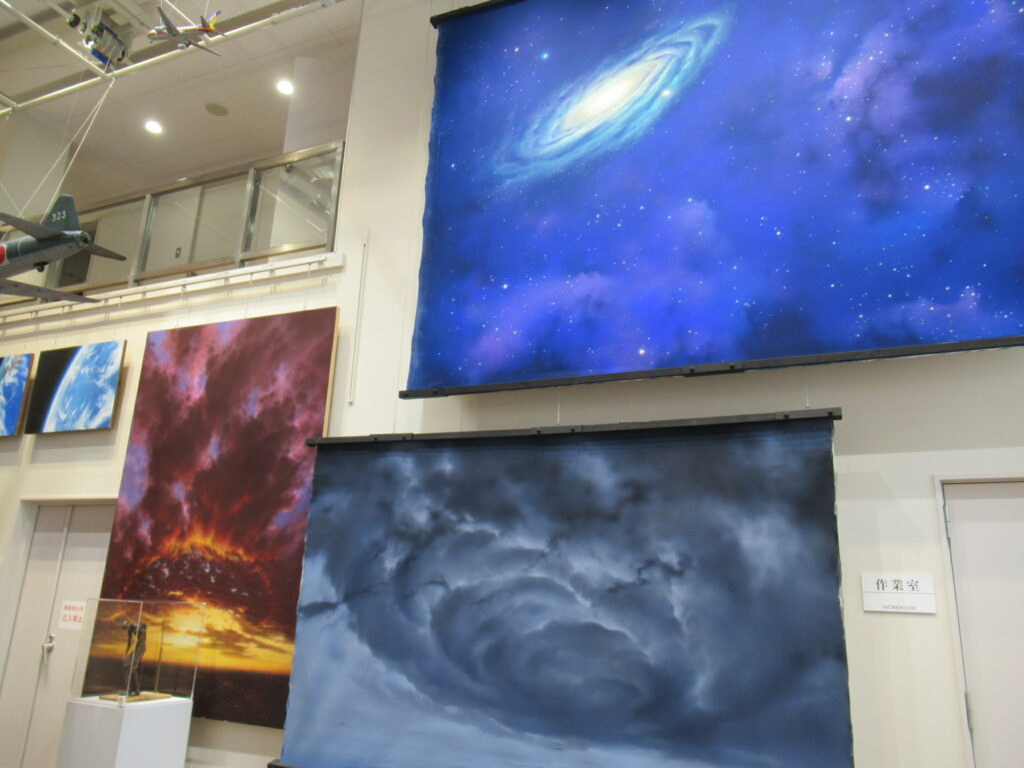 展示会場の白い壁面に、巨大な渦を巻く銀河が描かれた青い背景画や、不気味な暗雲が渦巻くグレーの背景画、燃えるような夕焼け空を描いた縦長の背景画が吊るされている写真