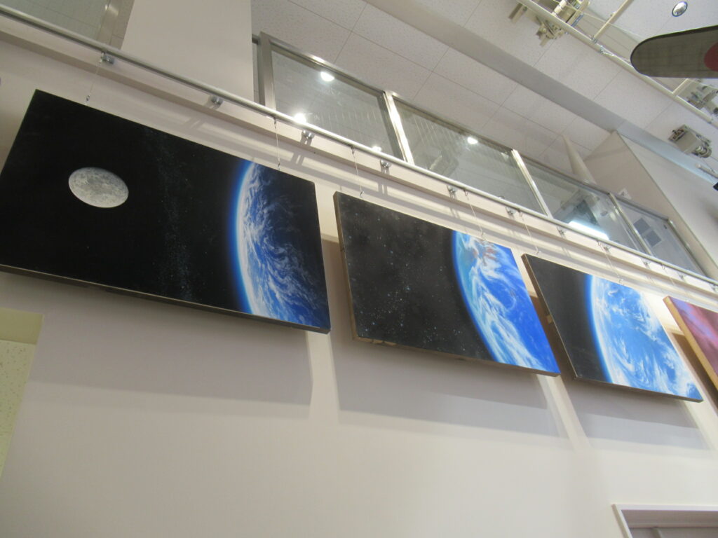 宇宙に浮かぶ輝く地球と月を描いた絵画が、高い天井を持つ白い壁面にワイヤーで吊るされており、下から見上げる角度で捉えた宇宙空間の背景画が3枚並んだ写真