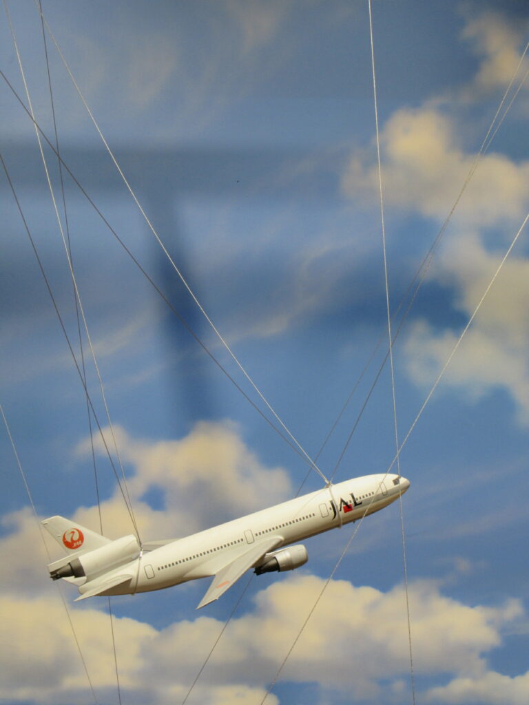 青空に白い雲が浮かぶ背景の前で、JALのロゴが入った白い旅客機の模型が多数の細いワイヤーによって吊るされ、機首を上げて力強く上昇しているような躍動感のあるディスプレイを捉えた写真