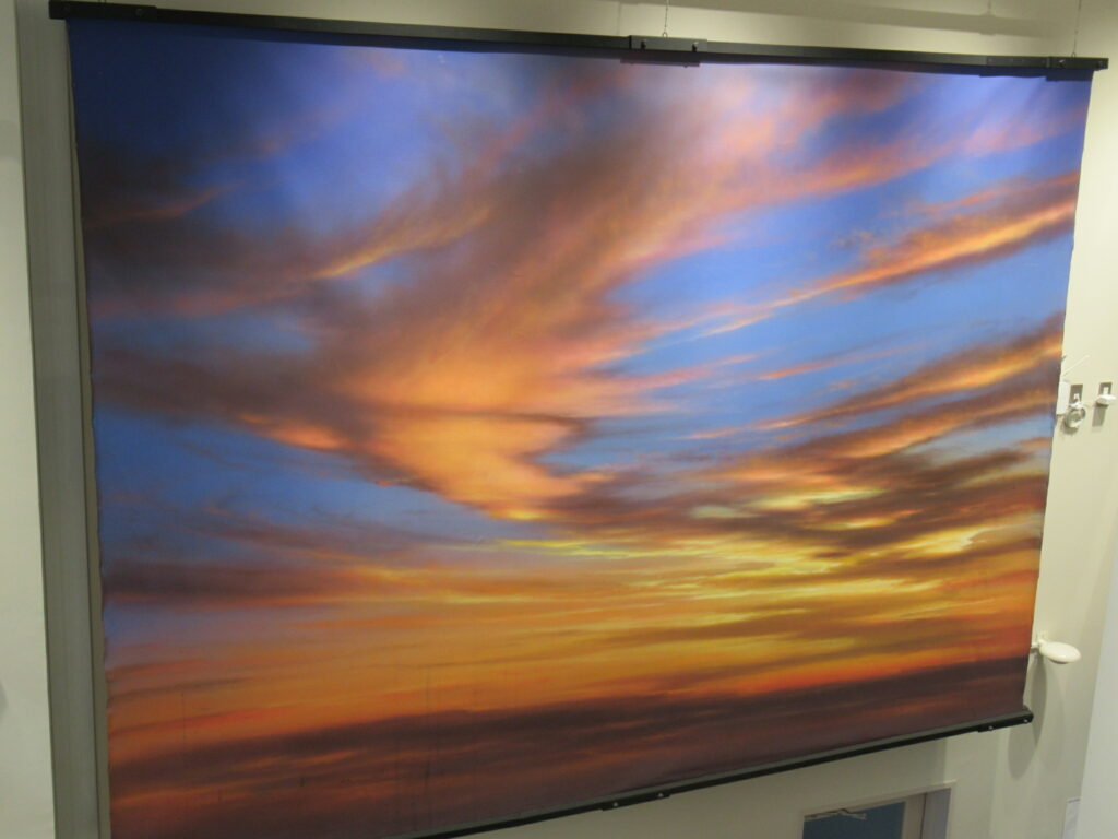 青色からオレンジ色へと美しく変化する夕焼け空が広がり、水平線に近い位置から放たれる黄金色の光がたなびく雲を下から照らし出している、壮大で叙情的な空の風景を豊かな色彩で描き出した大型の背景画の写真