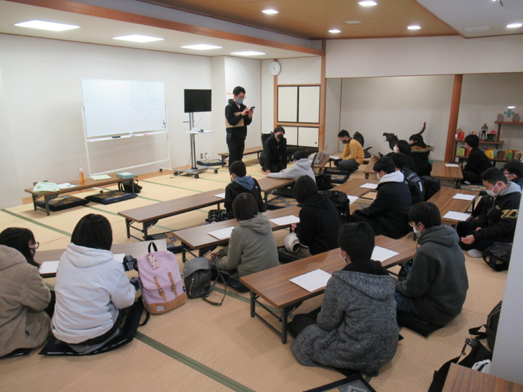 明るい和室の畳の上で、ホワイトボードやモニターを前にして、多くの塾生が低い長机を囲みながら座って説明を聞いたり資料を確認したりしている写真