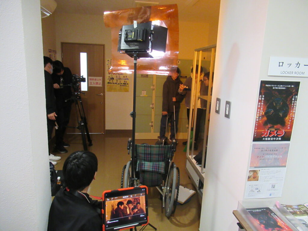 映画のポスターが貼られたロッカールーム前の通路で、車椅子に固定された照明機材や三脚付きのカメラを使い、モニターで映像を確認しながら真剣な表情で映画制作の撮影に臨んでいる塾生の活動風景の写真