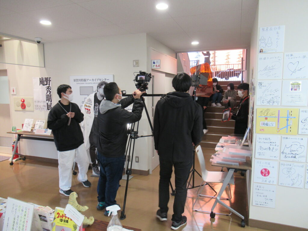 展示コーナーが並ぶ廊下にて、本格的な三脚に設置されたビデオカメラを構えるスタッフや、階段に座る出演者の様子を確認しながら、真剣な表情で撮影実習に取り組んでいる塾生の活動風景の写真