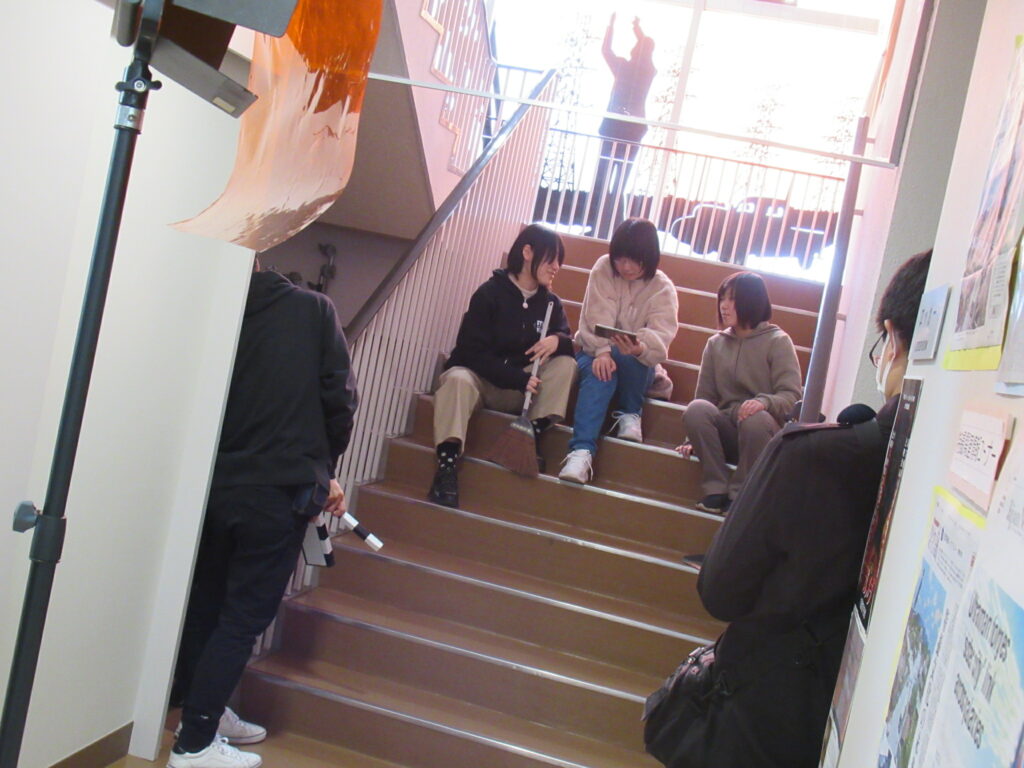 映画製作の現場にて、階段に座ってスマートフォンやタブレットを囲みながら談笑する3人の若者と、その様子を照明機材の影から見守るスタッフやカチンコを持つ人物を捉えた写真