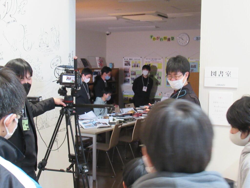 三脚に固定されたビデオカメラを操作して室内の様子を撮影している人物や、奥のテーブルに並べられた資料を囲んで作業の準備をするマスク姿の塾生を至近距離から写した写真