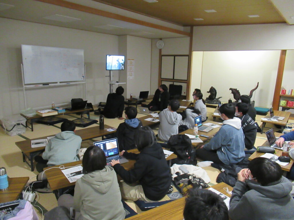 和室の部屋で、長机を囲んだ多くの塾生がノートパソコンを開き、正面に設置されたモニターを眺めながら映像を見ている写真