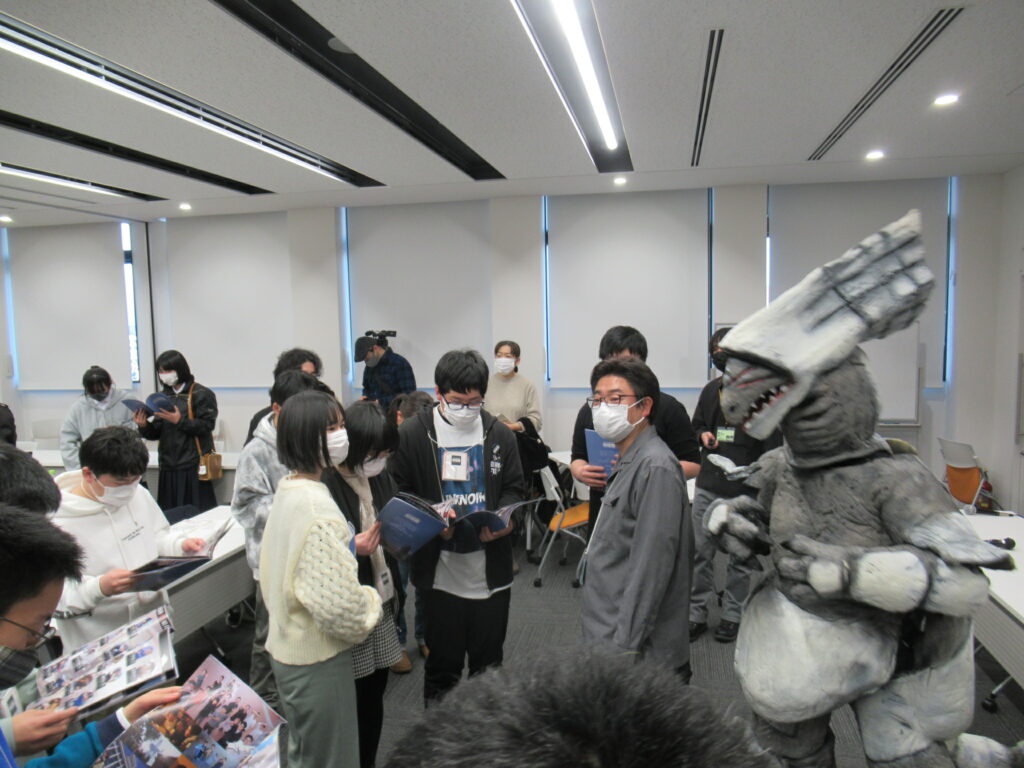 室内で特撮怪獣の着ぐるみが見守る中、多くの塾生たちが自分たちの活動がまとめられた冊子を手に取り、熱心に目を通している様子の写真